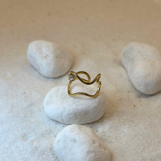 Un magnifique bague en acier inoxydable "Noeline" de couleur or, cette bagues scindé en deux, elle est de forme irrégulière avec un strass entre les deux formes, ce bijou est posé sur un caillou blanc qui est posé sur du sable blanc qui fait office de fond d'image.