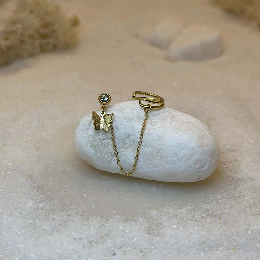 Un magnifique paire de boucle d'oreilles en acier inoxydable "Palma" de couleur Or, le bijou possède d'un cote un strass avec un papillon en pendentif ainsi qu'une bague de l'autre et est relié par une chaine, ce bijou est posé sur un caillou blanc qui  est posé sur du sable blanc qui fait office de fond d'image.