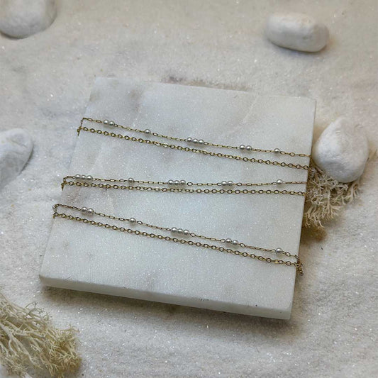 Une magnifique chaine de taille en acier inoxydable "Perla" de couleur or, ce bijou est posé sur une plaque de marbre blanche qui elle même est posé sur du sable blanc qui fait office de fond d'image.