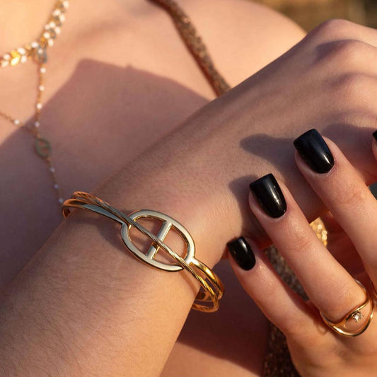 Une modèle porte de magnifiques bijoux en acier inoxydable de couleur doré / or. Nous y retrouvons le jonc "Romy" au design de noeud marin. En arrière plan se trouve les bagues "enea", "noeline", "ari", "inicia" et "mira". La lumière de la photo est chaude et nous y voyons le haut d'une robe dorée qui reflète le soleil.