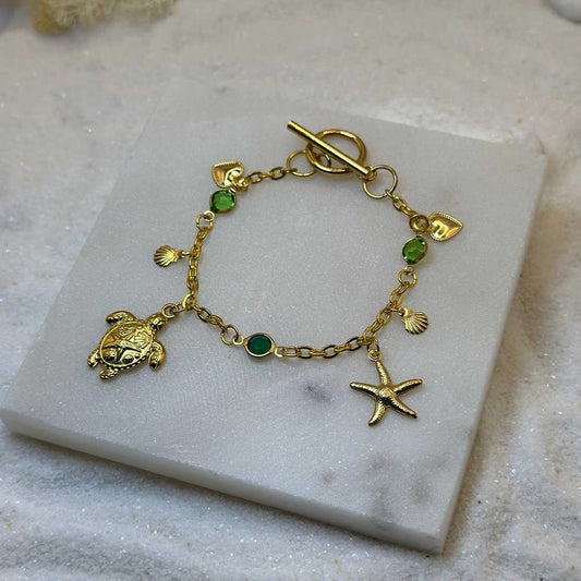 Un magnifique bracelet en acier inoxydable "Sania" de couleur or, ce bijou possède plusieurs charms : une étoile de mer, une tortue, un coquillage ainsi que deux coeurs, ci bijou est posé sur une plaque de marbre blanche qui elle même est posé sur du sable blanc qui fait office de fond d'image.