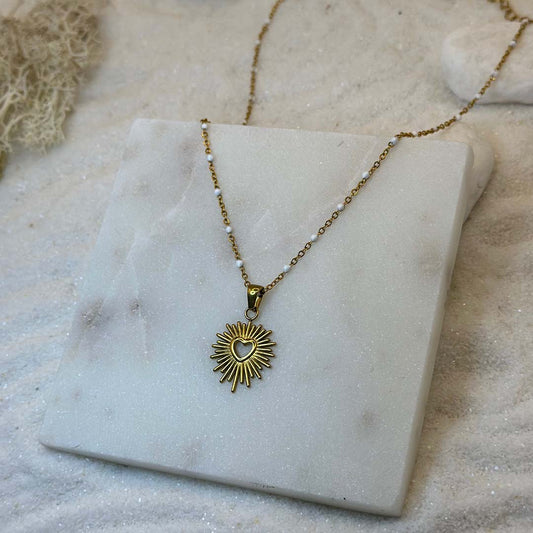 Un magnifique collier en acier inoxydable "Solia" de couleur Or, le bijou possède un pendentif de forme ronde avec un coeur au centre, ce bijou est posé sur une plaque de marbre blanche qui elle même est posé sur du sable blanc qui fait office de fond d'image.
