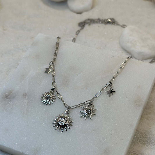 Un magnifique collier en acier inoxydable "Soline" de couleur argent, ce bijou possède plusieurs charms : 3 soleils de forme différentes ainsi qu'une étoile, le bijou est posé sur une plaque de marbre blanche qui elle même est posé sur du sable blanc qui fait office de fond d'image.