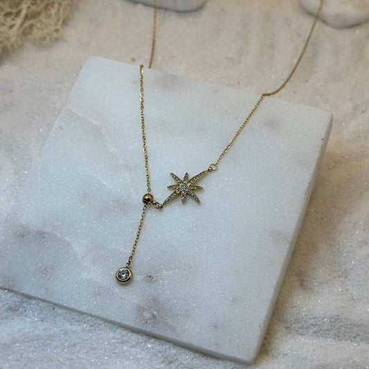 Un magnifique collier en acier inoxydable "Stella" de couleur or, ce bijou possède un charm en forme d'étoile incrusté de diamant ainsi qu'un strass en pendentif, ci bijou est posé sur une plaque de marbre blanche qui elle même est posé sur du sable blanc qui fait office de fond d'image.