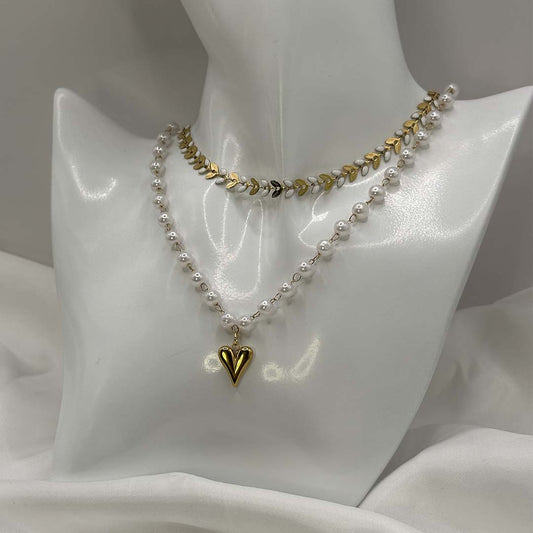 Deux magnifiques colliers en acier inoxydable sont posés sur un buste blanc brillant. Un collier "taina" est composé d'enchaînement de feuilles de couleur doré et blanc. Le 2e collier "Jane" est composé de perles blanches nacrées avec un pendentif en coeur de couleur or. C'est une composition imaginée par nos soins. Le buste est posé sur un fond blanc et un tissu en satin blanc. 