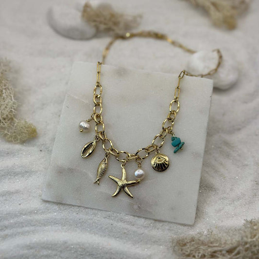 Un magnifique collier en acier inoxydable "Vaia" de couleur Or, le bijou possède plusieurs charms : deux perles blanches, deux coquillage, une étoile de mer et une pierre bleu turquoise, ce bijou est posé sur une plaque de marbre blanche qui elle même est posé sur du sable blanc qui fait office de fond d'image.