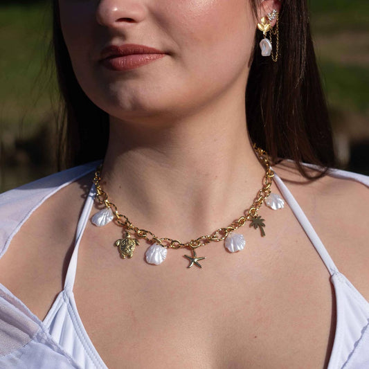 Un magnifique collier en acier inoxydable "Vanea" de couleur Or, le bijou possède plusieurs charms : plusieurs coquillages de couleur blanc, une tortue de couleur or, une étoile de mer de couleur or, un palmier de couleur or. Le collier est porté par une jolie modèle aux cheveux bruns. On y voit le bas de son visage ainsi que son oreille gauche avec les boucles "fidji" et "ezea". La modèle porte des vêtements blancs.