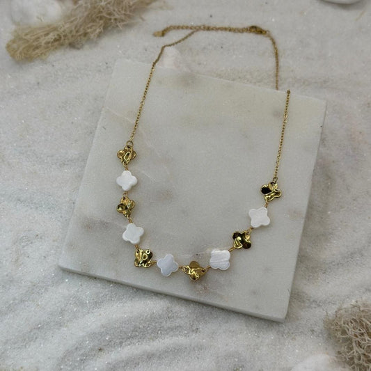 Un jolie collier en or "Inaya" est au centre de l'image, posé sur une plaque de marbre. Le bijoux en acier inoxydable est composé de trèfles or et blancs. Tout autour se trouve du sable blanc et des algues séchées.