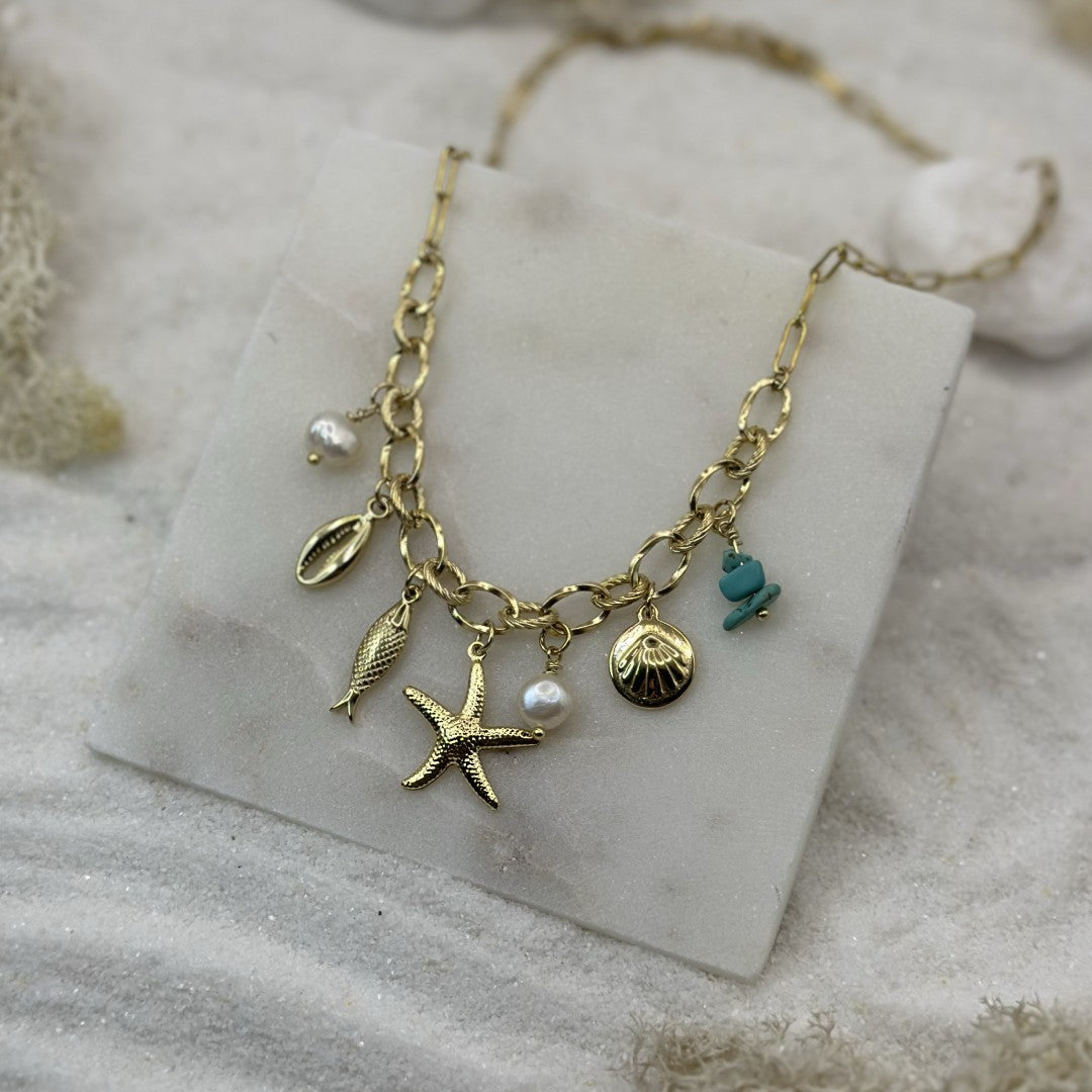 Un magnifique collier en acier inoxydable de la marque Atelier Vanéa est au centre de l'image. Le bijoux est couleur or avec différents charms : étoile de mer, coquillage, poisson et perle. Le bijoux est posé sur une plaque de marbre avec du sable autour.